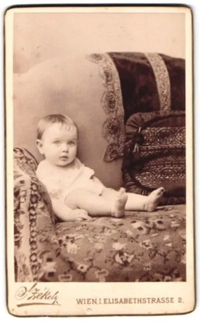 Fotografie Dr. Szekely, Wien, Elisabethstr. 2, niedliches Baby auf Sofa sitzend