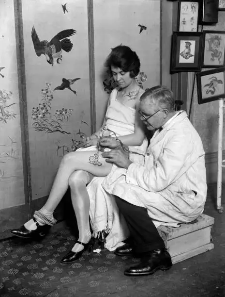 Woman having an image a snake tattooed onto her thigh by tattooist- 1930s Photo