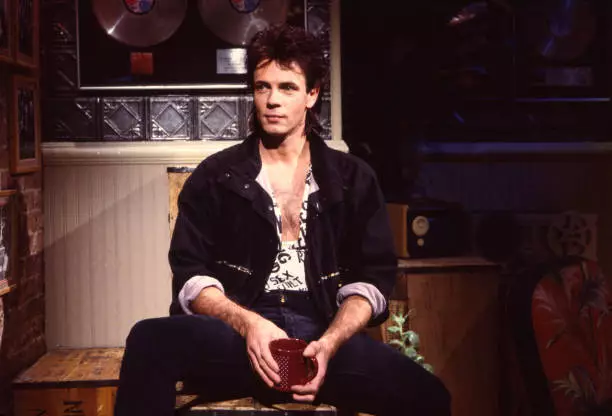 Rick Springfield as he sits on a crate, a coffee mug in his hands,  - Old Photo