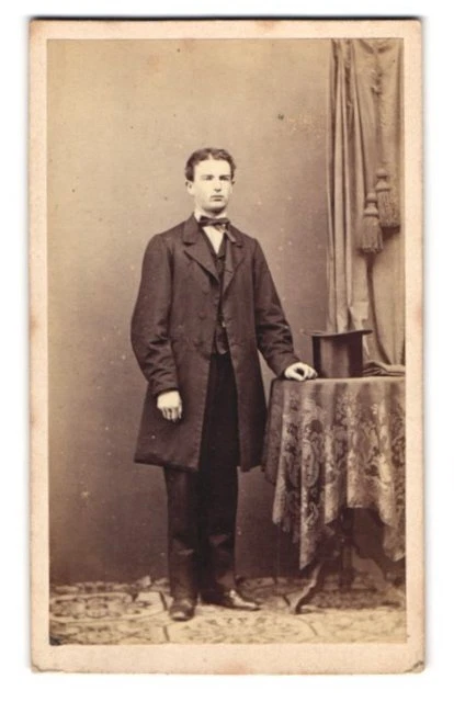 Fotografie W. Filser, Kaufbeuren, junger Mann im Anzug mit Zylinder auf dem Tis